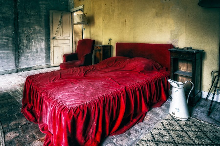 Run-down bedroom with a red velvet bed and armchair, dark wooden bedside furniture, a cream patterned rug and a white enamel pitcher, against mustard plaster and grey walls