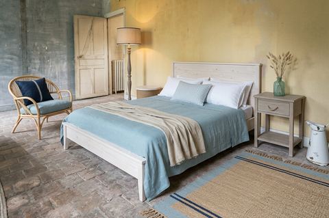 La même chambre en style bord de mer : lit en pin blanchi, couvre-lit en lin bleu mer, fauteuil en rotin avec coussin marine, chevet gris bois flotté et tapis en jute rayé