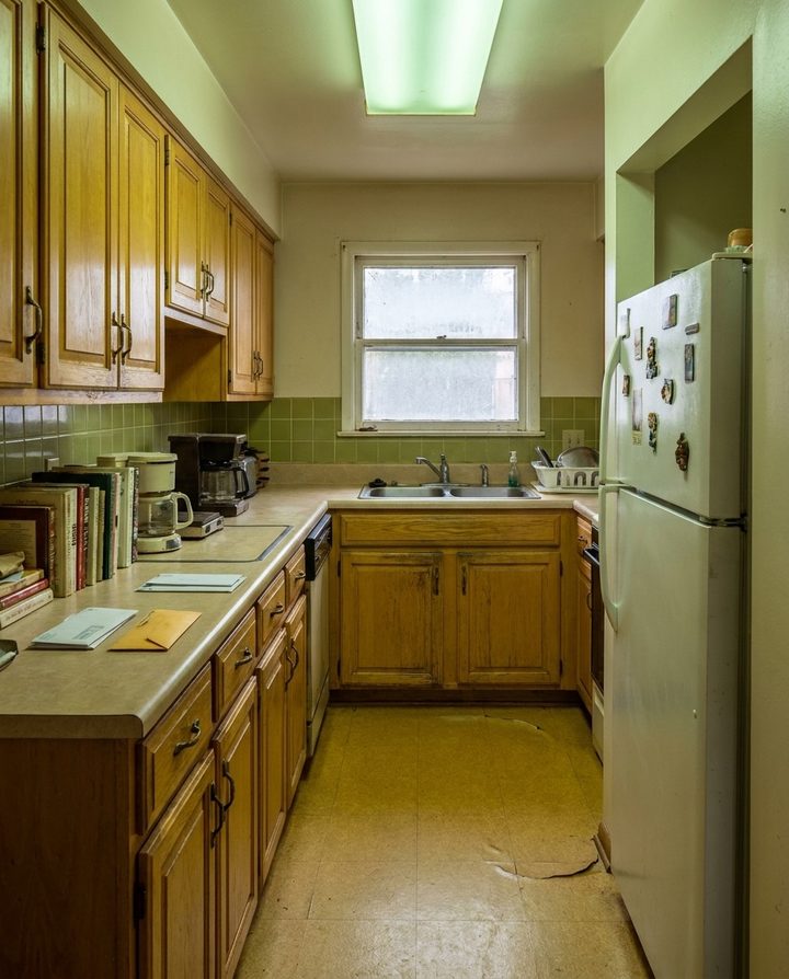 Cuisine couloir étroite avec meubles en chêne, plan stratifié beige, carrelage mural vert, évier double inox, plafonnier fluorescent, livres et papiers sur le plan et réfrigérateur blanc couvert d'aimants