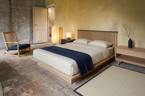 The same bedroom redesigned in Japanese style with a low pale oak platform bed, off-white linen, an indigo throw, an oak lounge chair, a paper lantern and a herringbone rush rug