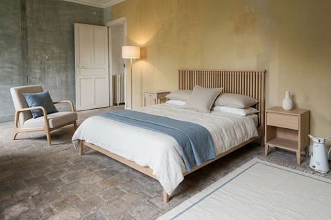 La même chambre en style scandinave : lit en chêne blond à tête de lit à lattes, linge de lit en lin blanc, plaid bleu poudré, fauteuil avoine et tapis clair tissé à plat