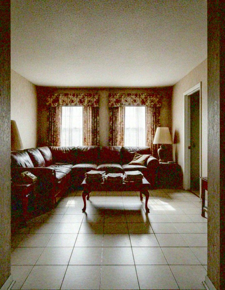 Dim living room with a dark brown leather L-shaped sectional, a cherry wood coffee table piled with magazines, dark side tables, a telephone, two table lamps and floral drapes with valances over two windows, on a beige tile floor.